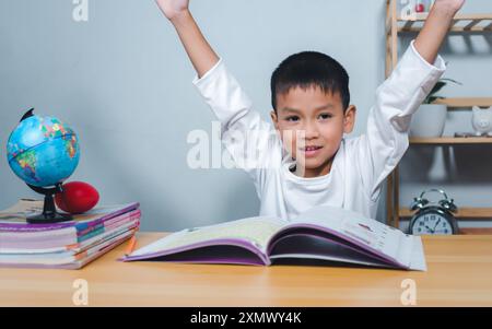 Concetto di istruzione, compiti e scuola. Bambino sorridente e alza felicemente la mano seduto a un tavolo con un libro. Bambini celebrati Foto Stock