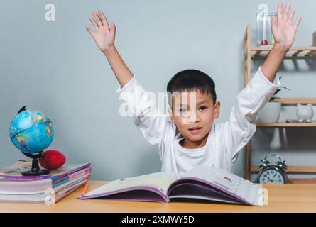 Concetto di istruzione, compiti e scuola. Bambino sorridente e alza felicemente la mano seduto a un tavolo con un libro. Bambini celebrati Foto Stock