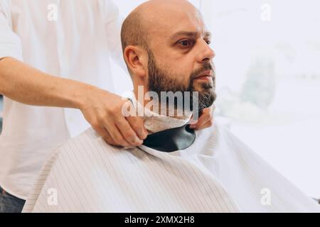 Facendo acconciature per uomo in un barbiere, l'uomo latino-americano fa un taglio di capelli a un uomo dalla barba calva nel salone del barbiere Foto Stock