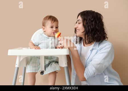 La mamma dava alla sua piccola bibita con il cibo in seggiolone su sfondo beige Foto Stock