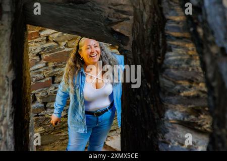 Donna turistica anziana che ride spontaneamente forte, capelli lunghi grigi, abiti casual, jeans e camicetta bianca, vista attraverso travi di legno, muri di pietra Foto Stock