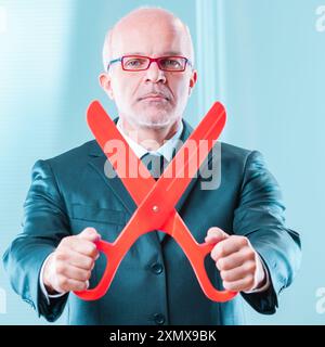 un uomo d'affari eterno con le forbici rosse rappresenta un duro taglio dei costi. La sua espressione risoluta sottolinea la necessità di ridurre i costi, le tasse, e. Foto Stock