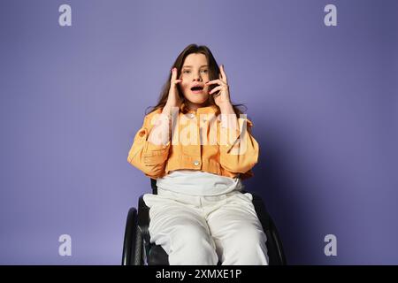 Giovane donna in un abbigliamento casual, in sedia a rotelle, siede in studio sullo sfondo viola, sorpreso dalle mani sopra la bocca Foto Stock