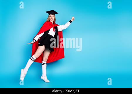 Foto di adorabile studentessa abbastanza positiva indossa un elegante abito rosso mantello alzare mano pugno volare spazio vuoto isolato su sfondo di colore blu Foto Stock