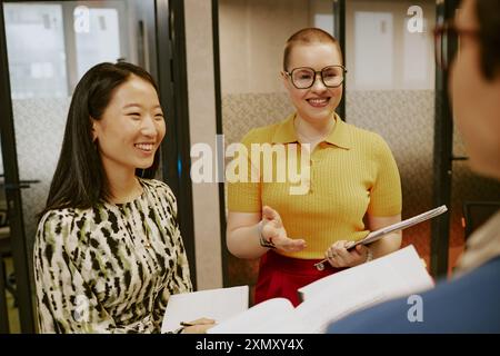 Due giovani manager asiatiche e caucasiche che detengono un quaderno e documenti in piedi in un ufficio moderno e parlano con la loro collega irriconoscibile Foto Stock