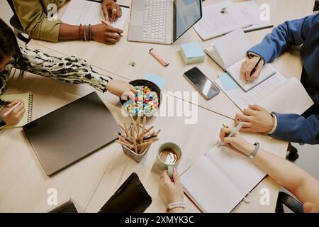 Immagine dall'alto di un gruppo di colleghi irriconoscibili seduti al tavolo e a prendere appunti, utilizzando un computer portatile e mangiando dolci Foto Stock