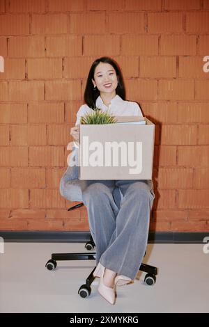 Colpo lungo verticale di giovane giovane asiatica allegra dipendente seduta su una sedia con una scatola in grembo contro un muro di mattoni rossi Foto Stock