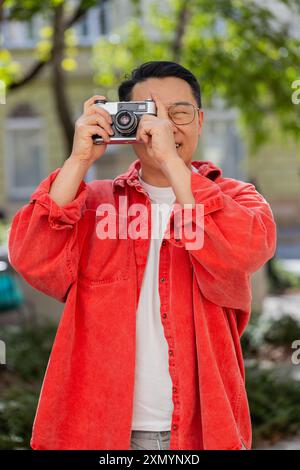 Uomo asiatico di mezza età turista che viaggia nei fine settimana esplorando la città, facendo foto con una macchina fotografica d'epoca retrò all'aperto. Un ragazzo cinese maturo in città. Verticale. Stili di vita Foto Stock