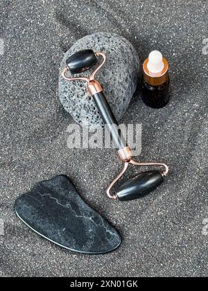 Strumenti per il massaggio facciale e olio essenziale su sabbia nera con sfondo pietra Foto Stock