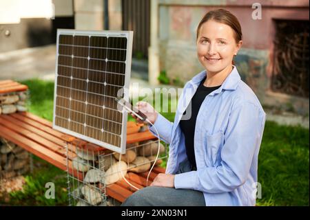 Donna felice che utilizza uno smartphone collegato al pannello solare fotovoltaico. Integrazione di energie rinnovabili sostenibili nella vita quotidiana, dimostrando l'uso pratico dell'energia solare per la ricarica dei dispositivi. Foto Stock