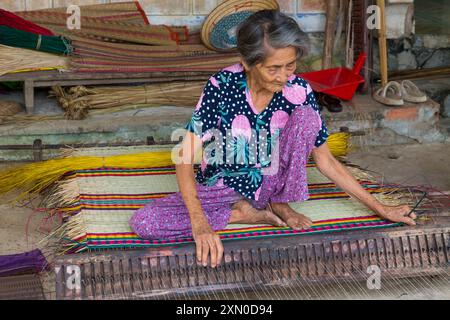 Anziana donna vietnamita che tesseva materassini da canne essiccate e tinte su telaio nel villaggio di tra Nhieu, Quảng Nam, Vietnam centrale, Asia a giugno Foto Stock