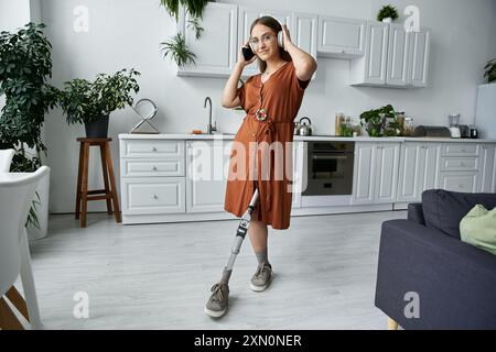 Una donna con un abito marrone e cuffie balla con una gamba protesica in una cucina luminosa. Foto Stock