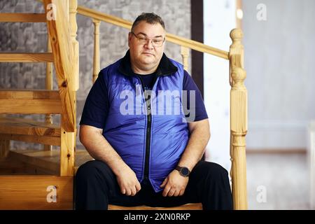 Uomo caucasico, 47 anni e sovrappeso, siede su gradini di scale di legno che conducono al secondo piano della sua casa. Foto Stock