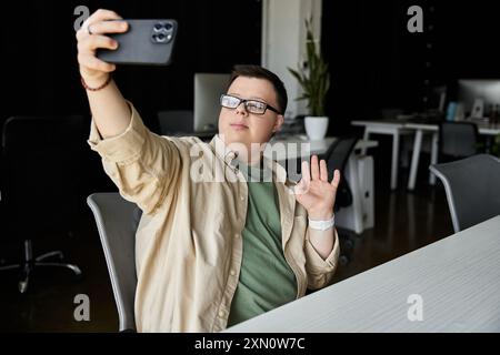 Un giovane con la sindrome di Down con gli occhiali sorride e onde mentre si fa un selfie in un ufficio. Foto Stock
