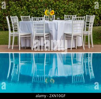 Tavolo preparato per cena, con sedie bianche di tovaglia e vaso con girasoli gialli, che si riflette sull'acqua blu della piscina Foto Stock