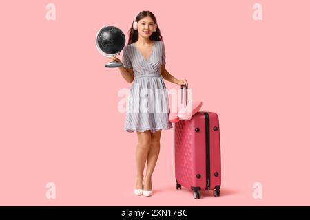 Giovane donna asiatica in cuffie con globo, cuscino da collo e valigia su sfondo rosa. Concetto di viaggio Foto Stock