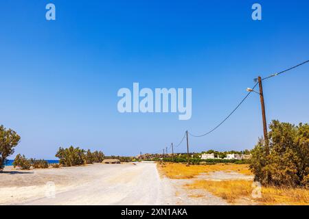 Strada sterrata lungo il mare con linee elettriche e pali che corrono lungo, circondata da un paesaggio asciutto con arbusti sotto un cielo azzurro. Creta. Grecia. Foto Stock