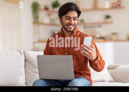 Uomo sorridente che lavora su un laptop e controlla il telefono in salotto Foto Stock