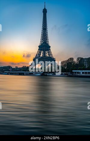 La Torre Eiffel si erge maestosamente contro un colorato cielo mattutino mentre il sole sorge, gettando riflessi sulle calme acque della Senna nelle vicinanze. Foto Stock