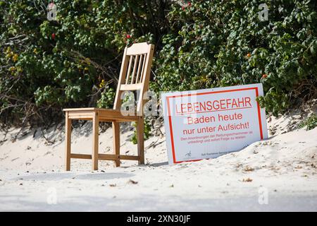 Strand von Westerland, Sylt - Baden verboten DEU, Deutschland, Sylt, Westerland: DAS Bild zeigt einen Holzstuhl, der neben einem Warnschild im Sand am Strand von Westerland auf Sylt steht. DAS Schild trägt die Aufschrift LEBENSGEFAHR Baden heute nur unter Aufsicht Den Anordnungen der Rettungsschwimmer ist unbed und sofort Folge zu leisten. Im Hintergrund sind dichte Büsche und Pflanzen zu sehen, die dem Strand eine natürliche Grenze setzen. Westerland Sylt Schleswig Holstein Deutschland *** Spiaggia di Westerland, Sylt Swimming Prohibited DEU, Germania, Sylt, Westerland l'immagine mostra un woo Foto Stock