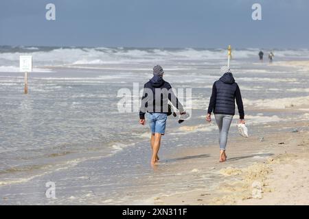 Spaziergang am stürmischen Strand von Westerland, Sylt DEU, Deutschland, Sylt, Westerland: DAS Bild zeigt zwei Personen, die entlang des Strandes von Westerland auf Sylt spazieren. SIE tragen Jacken und Mützen und halten ihre Schuhe in den Händen, während sie barfuß im Sand laufen. Die Nordsee zeigt sich mit starken Wellen und der Himmel ist bewölkt. Westerland Sylt Schleswig Holstein Deutschland *** camminate sulla spiaggia tempestosa di Westerland, Sylt DEU, Germania, Sylt, Westerland la foto mostra due persone che camminano lungo la spiaggia di Westerland su Sylt indossano giacche e berretti e tengono in mano il loro sho Foto Stock