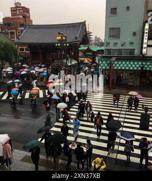 Ombrelli e persone che attraversano una trafficata strada di Tokyo sotto la pioggia, creando una vivace scena urbana. Foto Stock