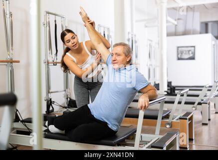 Giovane istruttrice positiva che controlla i movimenti dell'uomo anziano che si allunga sulle attrezzature del riformatore Pilates in studio Foto Stock