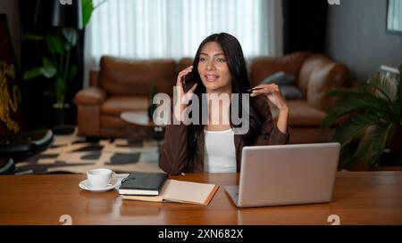 Una donna d'affari asiatica sicura e intelligente, con la pelle abbronzata, indossa un abito marrone, sta parlando al telefono con un cliente a una scrivania nella sua moderna metà del secolo Foto Stock