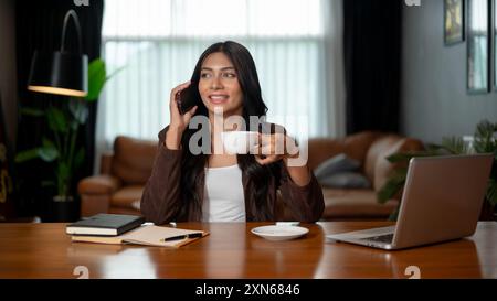 Una donna d'affari asiatica sicura e intelligente, con la pelle abbronzata, indossa un abito marrone, sorseggia un caffè mentre parla al telefono con un cliente a una scrivania Foto Stock