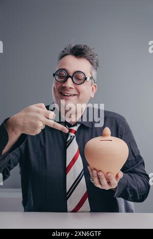 L'uomo d'affari che indossa occhiali spessi e divertenti indica un salvadanaio che tiene con sé con un'espressione allegra e sciocca Foto Stock