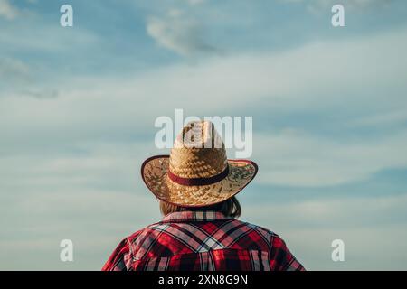 Vista posteriore di una donna che indossa un cappello di paglia in piedi nel campo e guarda l'orizzonte sulle colture pronte per il raccolto in una soleggiata giornata estiva, selectiv Foto Stock