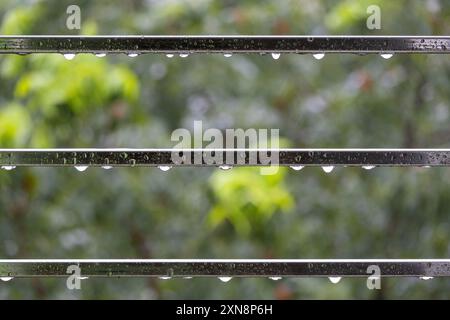 L'acqua piovana gocciola sul tubo in acciaio nelle giornate piovose con sfondo verde sfocato e luce bokeh. La pioggia cade sul tubo d'acciaio del balcone. Foto Stock