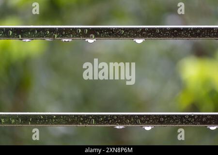 La pioggia cade sul tubo d'acciaio del balcone. L'acqua piovana gocciola sul tubo in acciaio nelle giornate piovose con sfondo verde sfocato e luce bokeh. Foto Stock