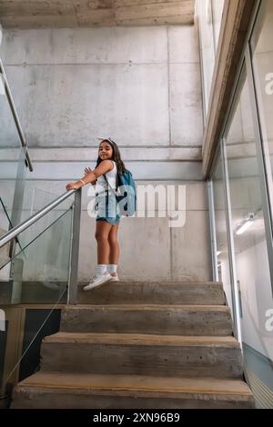 Una ragazza con una maglietta bianca e pantaloncini blu è in piedi su una scala che entra nella scuola. Indossa uno zaino e calzini bianchi. L'immagine ha una c Foto Stock