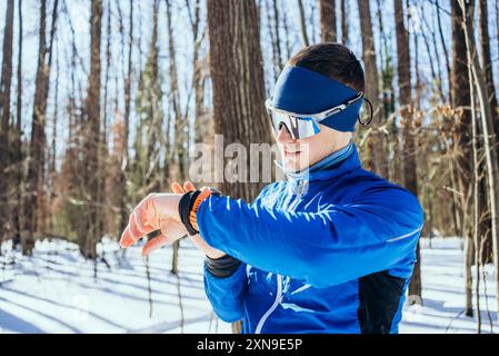 L'atleta con gli outfit invernali blu controlla il suo fitness tracker mentre corre in una foresta innevata, mostrando sport invernali e fitness. Uomo su treni sci di fondo nella foresta invernale. Foto Stock