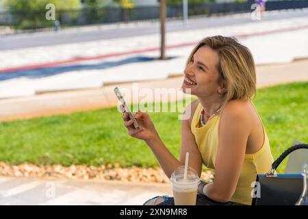 Una giovane donna sorridente ama il suo caffè ghiacciato mentre controlla il telefono in un ambiente esterno. Lo sfondo vivace della città e il suo stile casual riflettono un Foto Stock