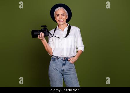 Foto dell'adorabile e adorabile camerawoman indossa camicia bianca cappello per registrare video macchina fotografica professionale spazio vuoto sfondo verde isolato Foto Stock