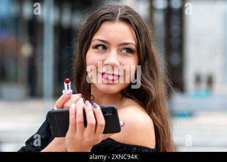 Una donna sorride mentre applica il rossetto rosso mentre tiene il telefono. Foto Stock