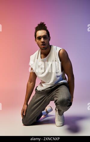 Un giovane afroamericano con una camicia bianca senza maniche e pantaloni grigi posa su un ginocchio su uno sfondo colorato e brillante. Foto Stock