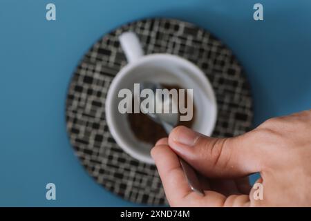 Mano di un uomo bianco che versa caffè istantaneo in una piccola tazza bianca su un tavolo da cucina blu per la colazione. Foto Stock