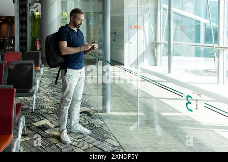 Uomo con zaino che usa il telefono in piedi contro la finestra dell'aeroporto Foto Stock