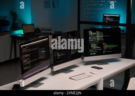 Immagine di sfondo dell'ufficio di sviluppo IT e tre monitor di computer sul tavolo in luce blu, spazio di copia Foto Stock
