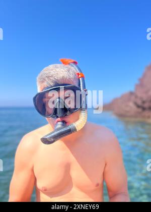 Un uomo sta guardando la macchina fotografica, indossando l'attrezzatura da snorkeling e occhiali di protezione, pronto a tuffarsi in acqua. Lo sfondo mostra cieli azzurri e scogliere rocciose Foto Stock