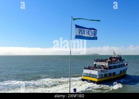 Molo 39 bandiera del Fisherman's Wharf che fluttua sopra un traghetto Osky della compagnia Blue and Gold Fleet. Foggy Alcatraz Island sullo sfondo - San fra Foto Stock