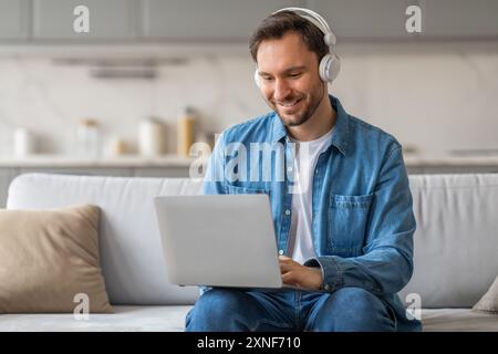 L'uomo sorridente utilizza il laptop mentre indossa le cuffie su un divano Foto Stock