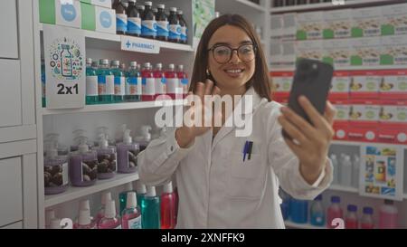 Una giovane, bella, farmacista con un camice bianco, capelli e occhiali bruna, che fa una videochiamata all'interno di una farmacia ben fornita, sorridendo Foto Stock