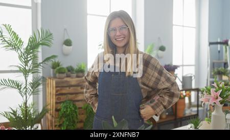 Una giovane donna allegra con camicia in flanella e grembiule in denim si erge tra le piante in vaso all'interno di un negozio di fiori illuminato dal sole. Foto Stock