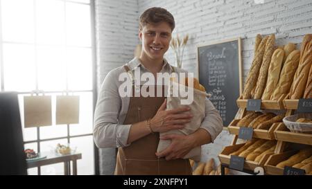 Handome giovane uomo in una panetteria che tiene le baguette appena sfornate in un sacco di stoffa, sorridendo sullo sfondo di scaffali del pane e un tabellone da menu in un pozzo Foto Stock