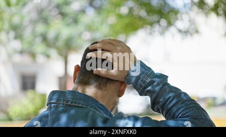 Vista posteriore di un uomo latino calvo e barbuto all'aperto, che tiene la testa in un'atmosfera contemplativa in un verde parco cittadino. Foto Stock
