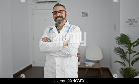 Medico ispanico maturo e sicuro con barba grigia braccia in piedi attraversate nel corridoio della clinica, che ritrae professionalità ed esperienza. Foto Stock